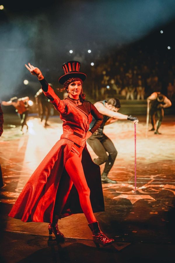 “Let’s Get Wicked” actress Annabel Forman, seen in a red dress and top hat, raises her arm in performance in front of background dancers.