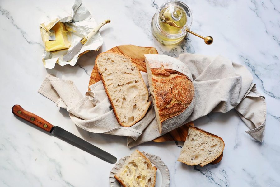A sourdough loaf sits on a cloth on top of a marble counter, surrounded by bread slices, a knife, honey and butter.
