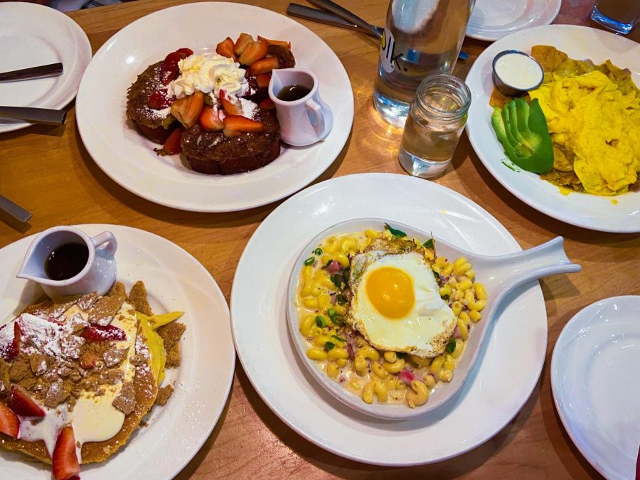 On a brown wooden table sits white plates holding Strawberry Cheese Cakes, Red Velvet French Toast, Breakfast Mac and Cheese and Chilaquiles Verdes.