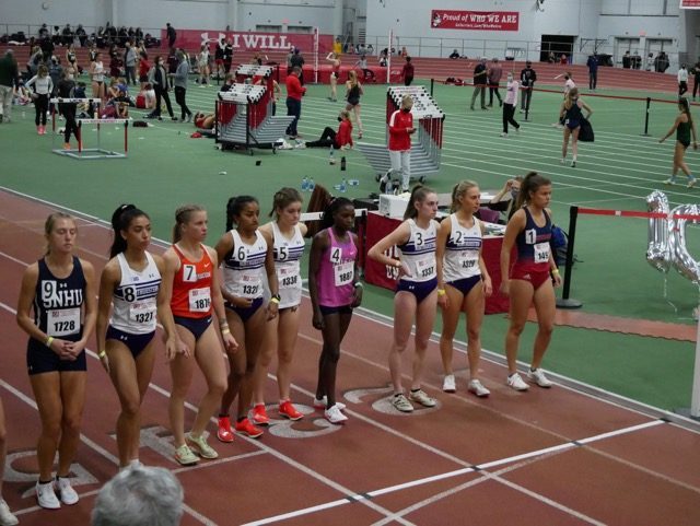 A group of female runners line up before the start of a race on an indoor track.