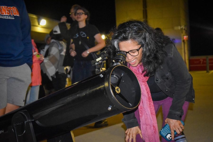 A person wearing purple pants, a black jacket, a pink scarf and glasses looks through the eyepiece of a black telescope in front of a crowd.