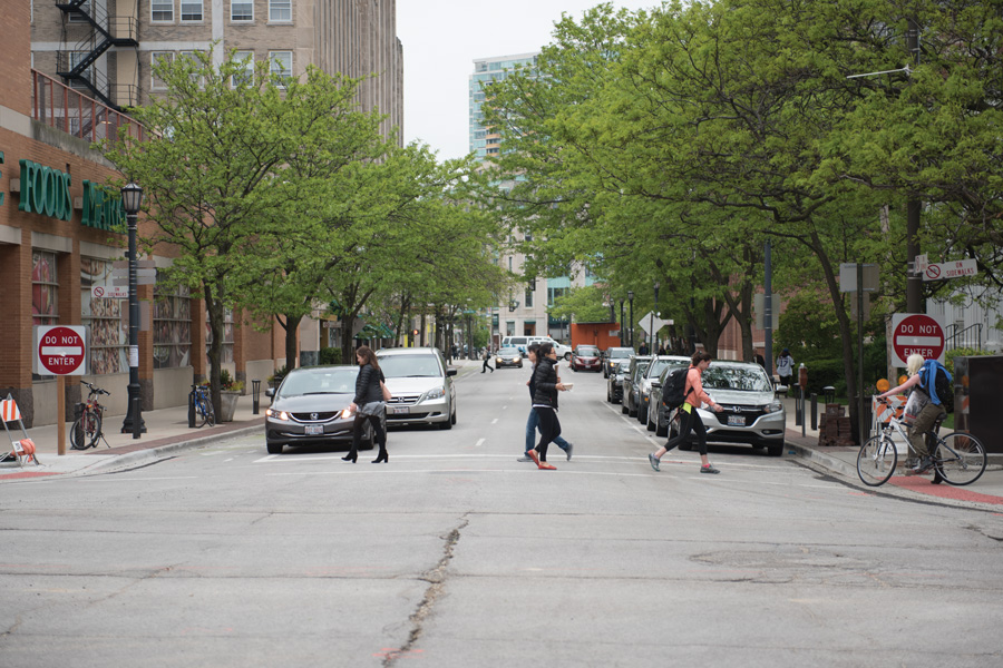 People cross a street in downtown Evanston. The crosswalk, located next to a Whole Foods, is lined with trees.