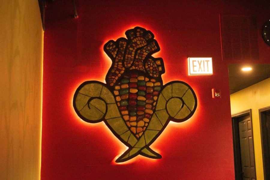 A sculpture of an ear of corn shaped like a heart hangs on a red wall.