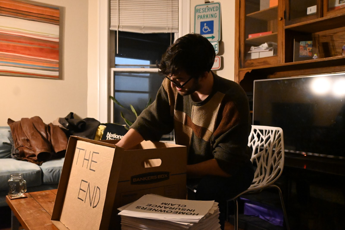 A person sits in a living room and reaches into a cardboard box next to a thick stack of papers.